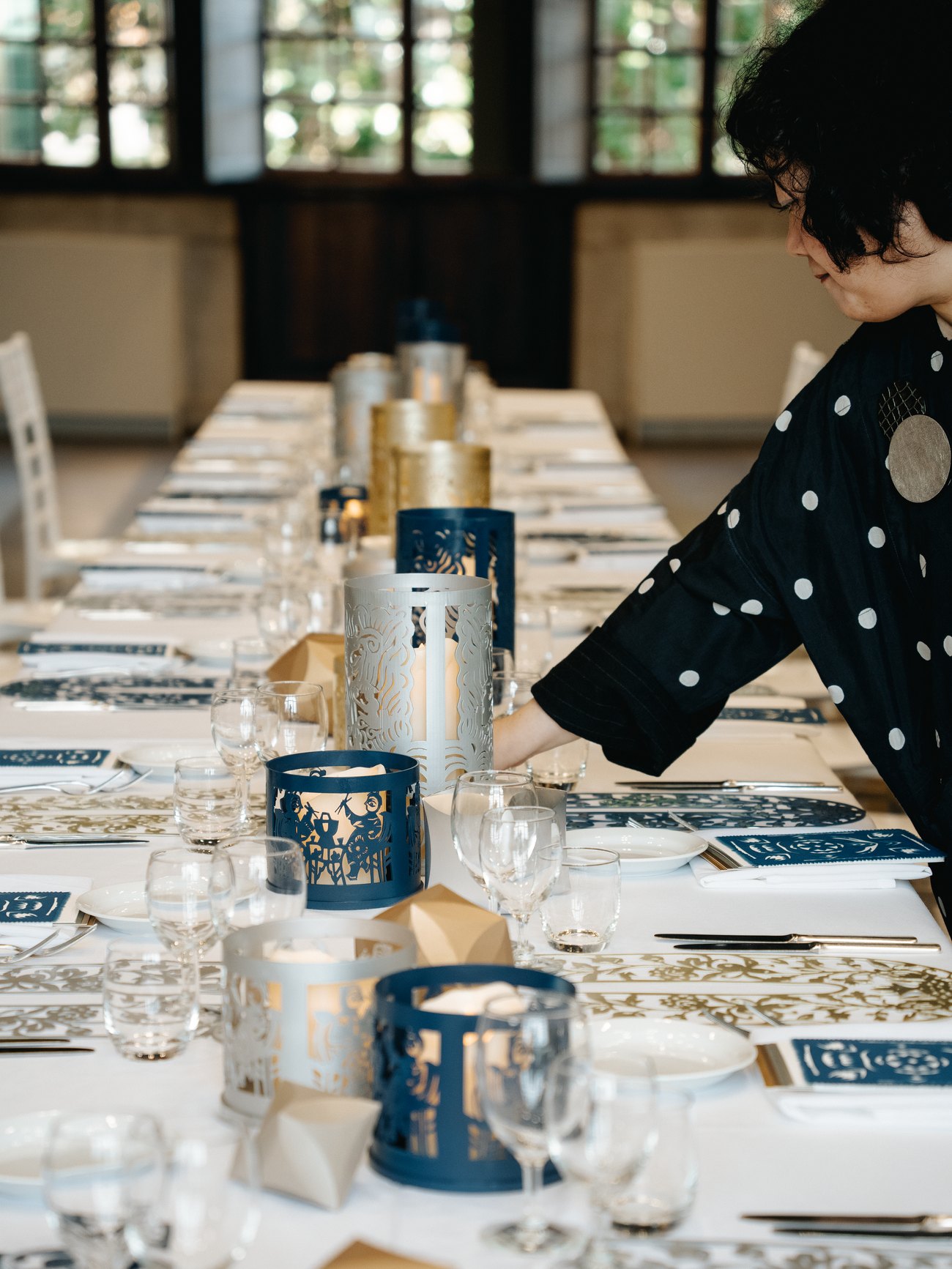 A woman in a black polka-dot blouse leans across a long banquet table, placing pierced paper-cut lanterns among the place settings while the room is still bare of guests.