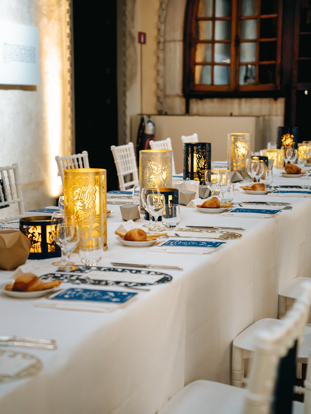 The Founding Dinner table at dusk: pierced lanterns glowing gold along a long white cloth, with bread, blue menu cards and cutlery laid for guests.
