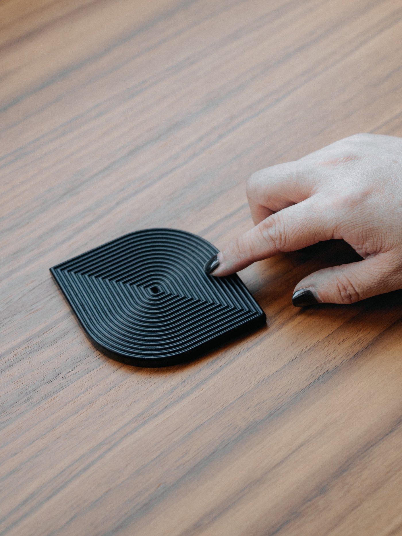 A hand resting on The Eye of Wisdom: a black 3D-printed table piece whose concentric square ridges turn inward towards a small square void at the centre.