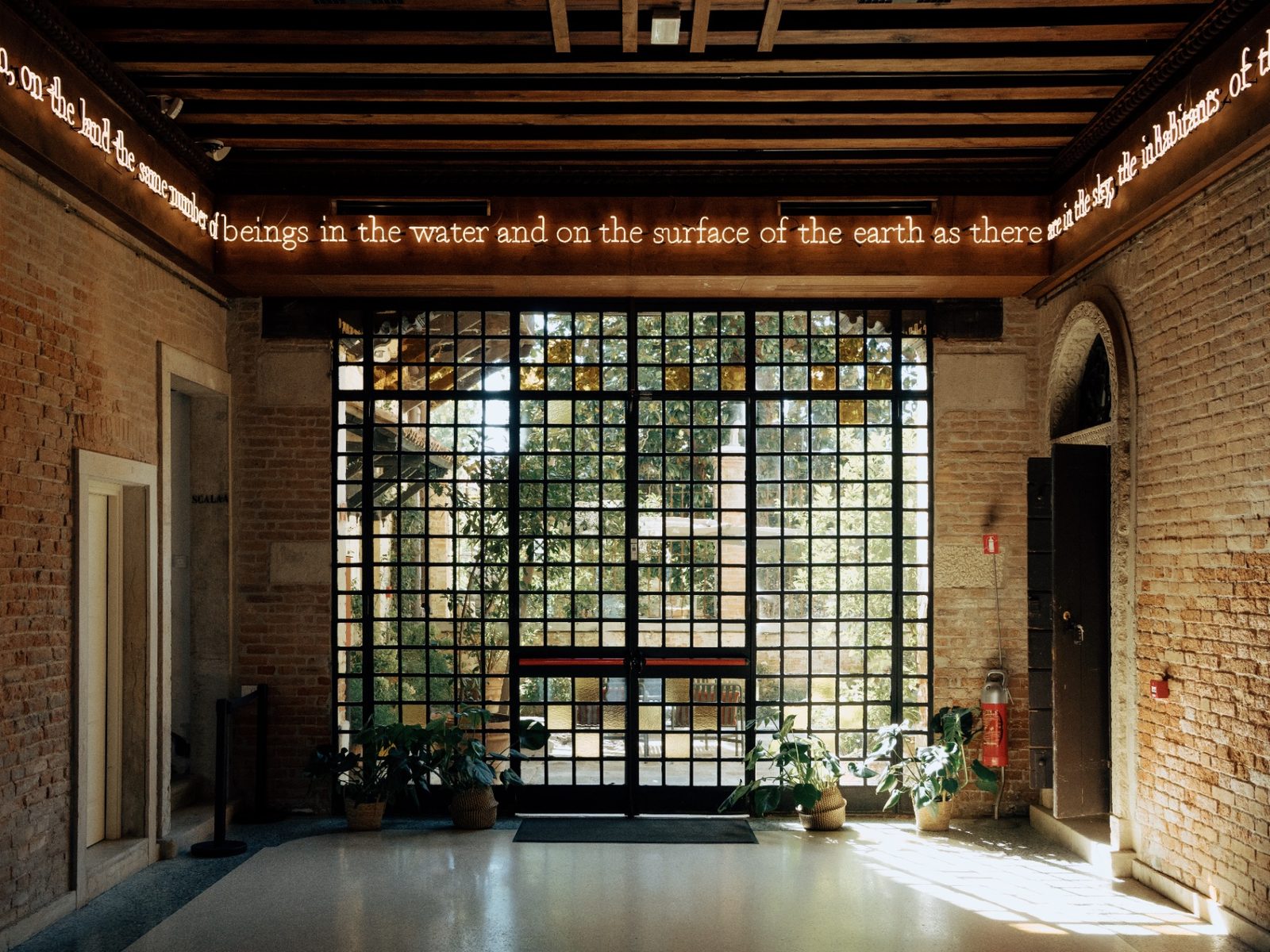 The entrance foyer of Casa dei Tre Oci: brick walls, a grid-glass door opening toward the Giudecca canal, and a neon text installation running along the ceiling.