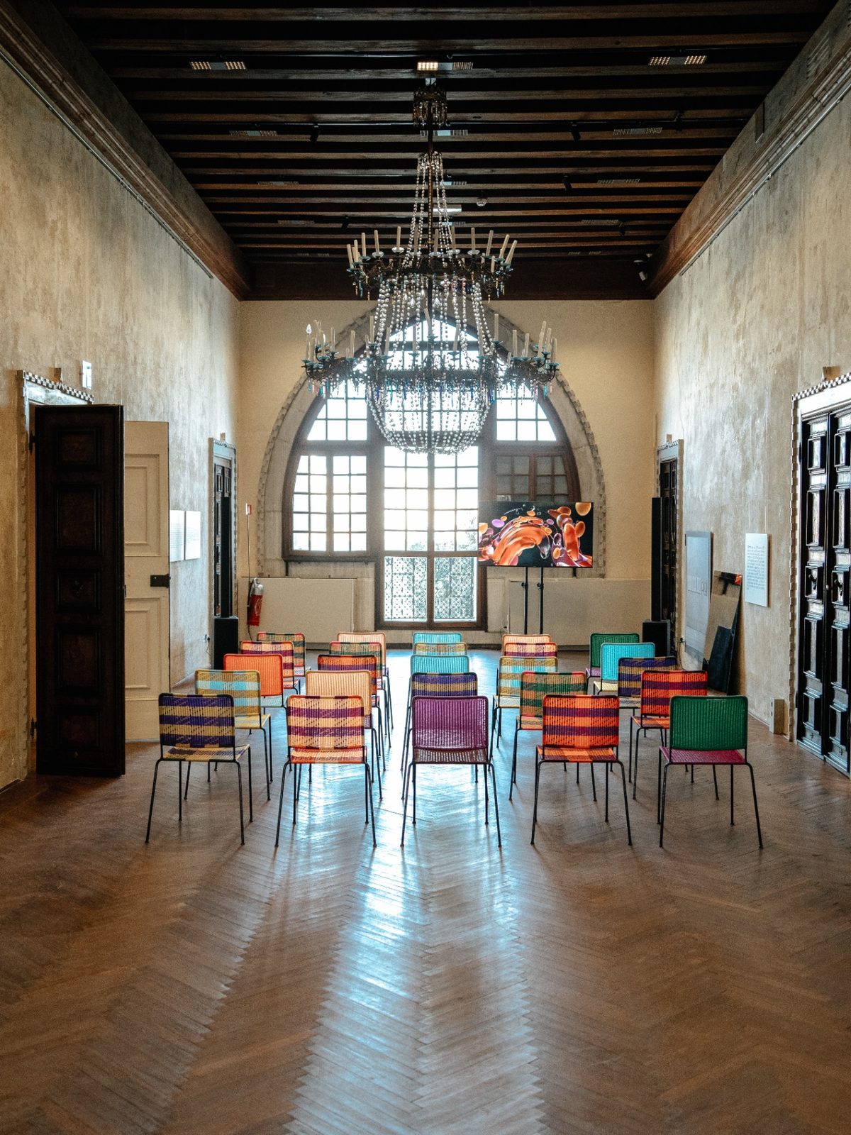 The main hall of Casa dei Tre Oci: a Venetian glass chandelier, a herringbone parquet floor and colourful woven chairs arranged for a conversation.